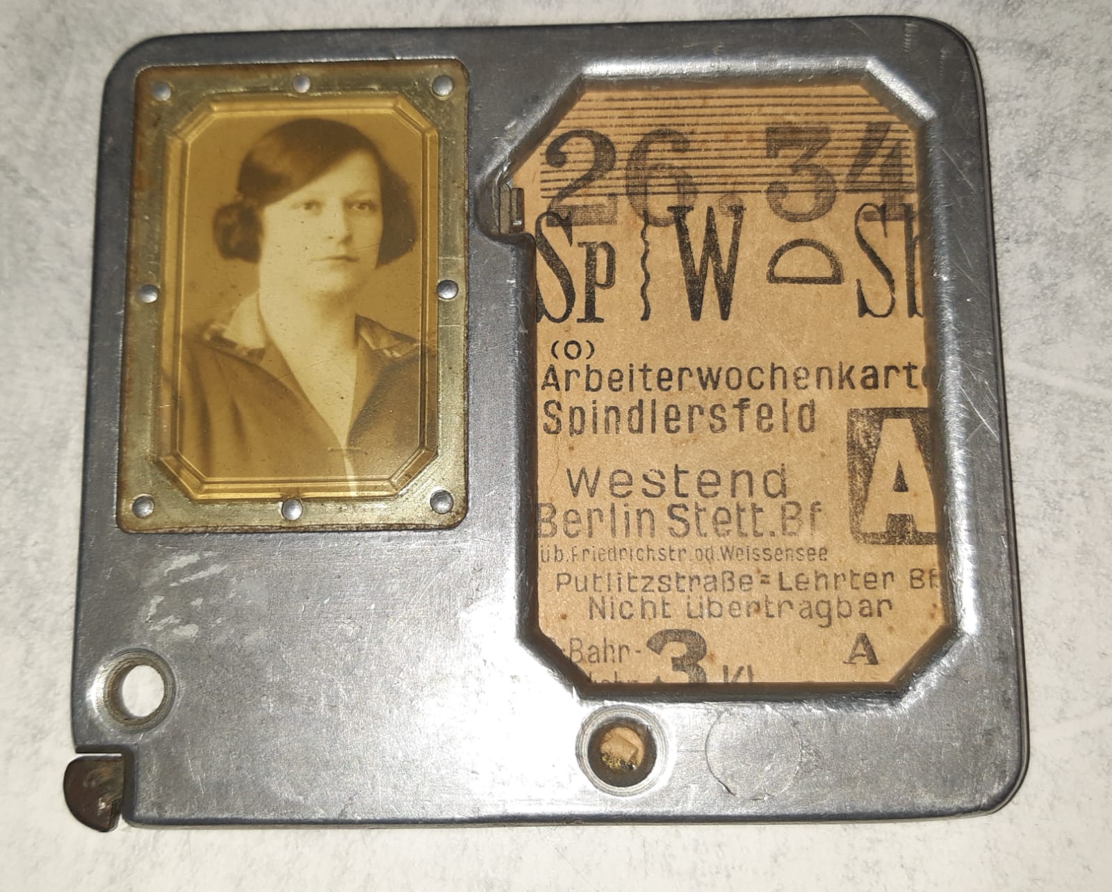 Ein quadratischer Metallrahmen mit links einem s/w-Bild einer jungen Frau und daneben, rechts, einer Pappfahrkarte für die 26. Kalenderwoche des Jahres 1934. Sie gilt laut Aufdruck für die 3. Klasse von Spindlersfeld nach Westend.